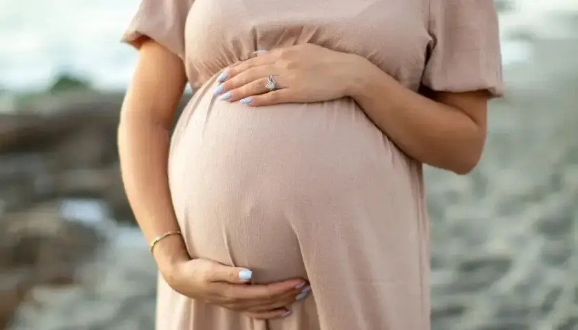 Brzuch kobiety w ciąży, delikatnie otulony beżową sukienką. Dłonie z jasnoniebieskim lakierem na paznokciach spoczywają na brzuchu. Warto pamiętać o bezpieczeństwie, np. unikając retinolu w ciąży.
