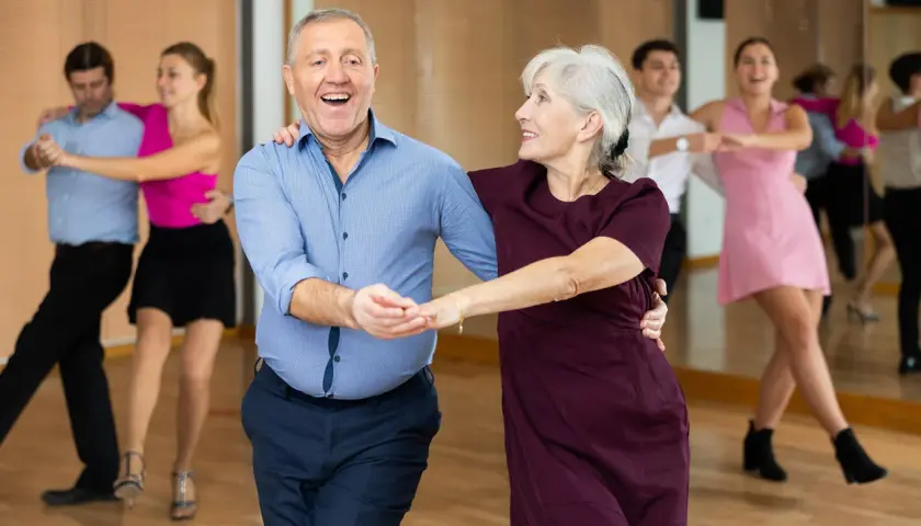 Uśmiechnięta para seniorów w rytmie tańca na 3. W tle inne pary ćwiczą kroki.