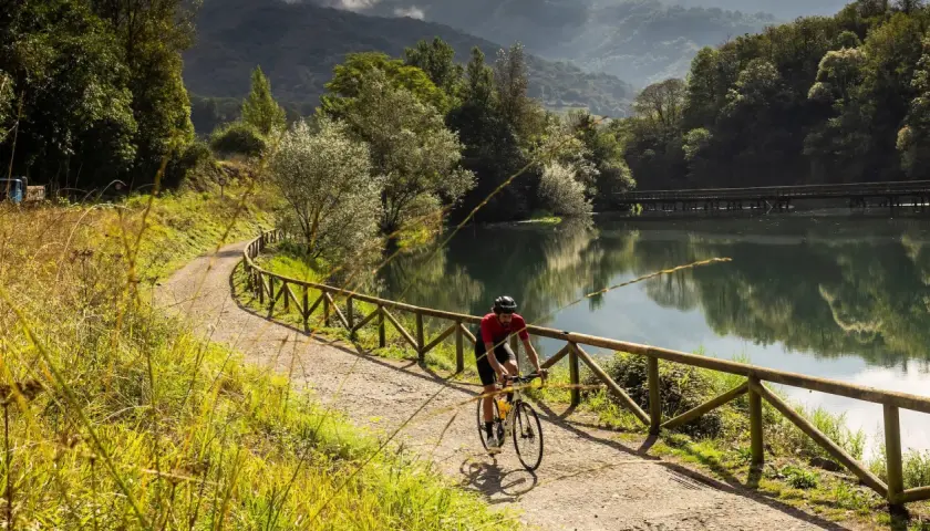Ciclista pedalea por una de las vías verdes España, junto a un lago sereno y rodeado de exuberante vegetación.