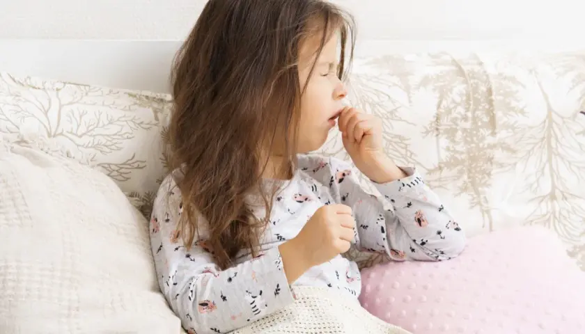 Bambina con tosse stizzosa, coperta da una coperta, seduta a letto con cuscini decorati.