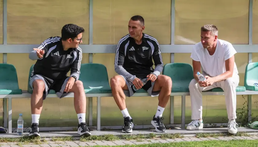 Trzech mężczyzn siedzi na ławce. Dwóch w strojach sportowych, jeden w białej koszulce. To trener Lechii Gdańsk i jego współpracownicy.