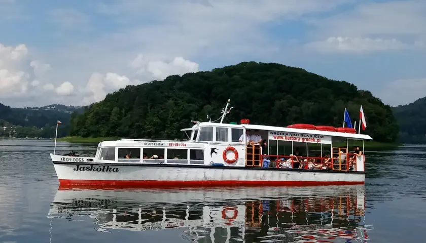 Statek "Jaskółka" pływa po zalewie rożnowskim, wioząc turystów wśród zielonych wzgórz.