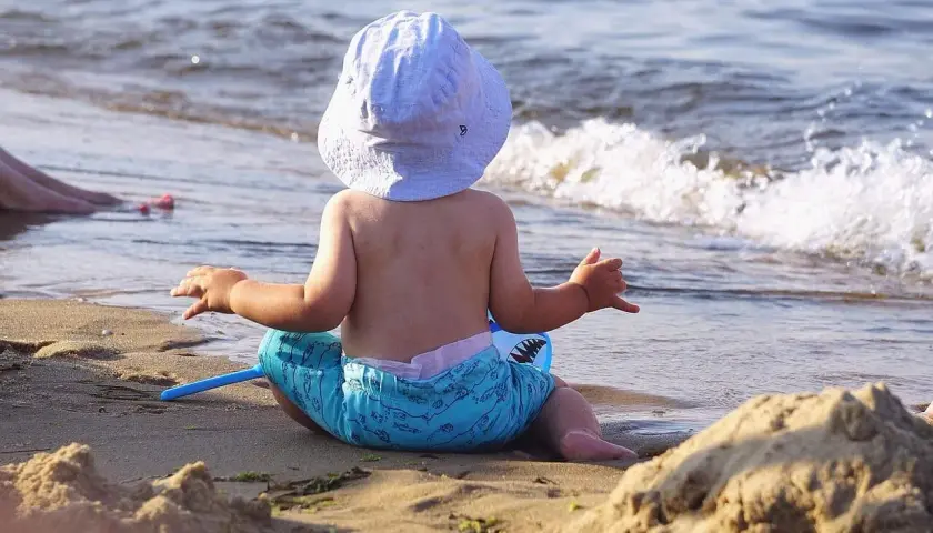 Niemowlę w kapeluszu i kąpielówkach z rekinami siedzi na plaży, ciesząc się wakacjami z niemowlakiem nad morzem.