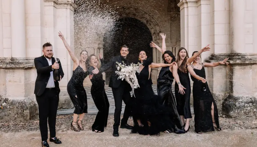 Un groupe joyeux célèbre un mariage noir et blanc, le marié aspergeant de champagne ses demoiselles d'honneur en robe noire.