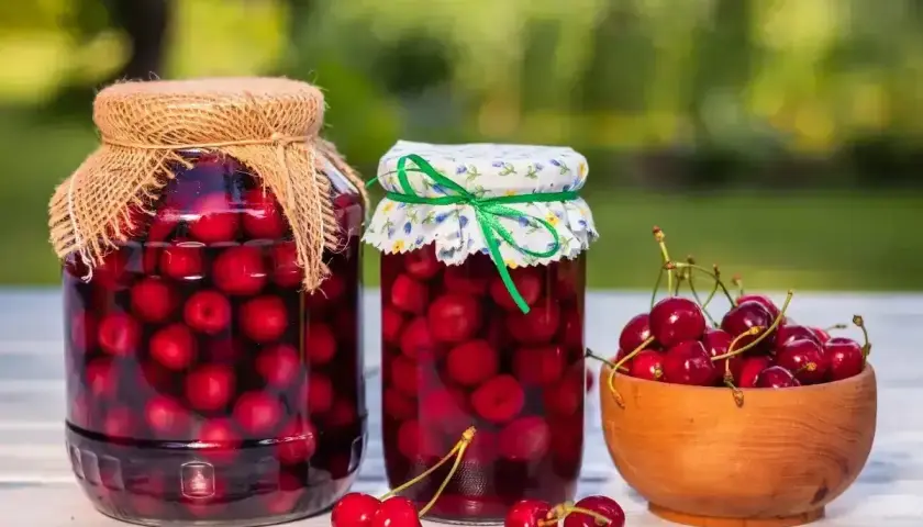 Dwa słoje domowego kompotu z czereśni, jeden z lnianym, drugi z kwiecistym nakryciem. Obok miseczka świeżych czereśni.