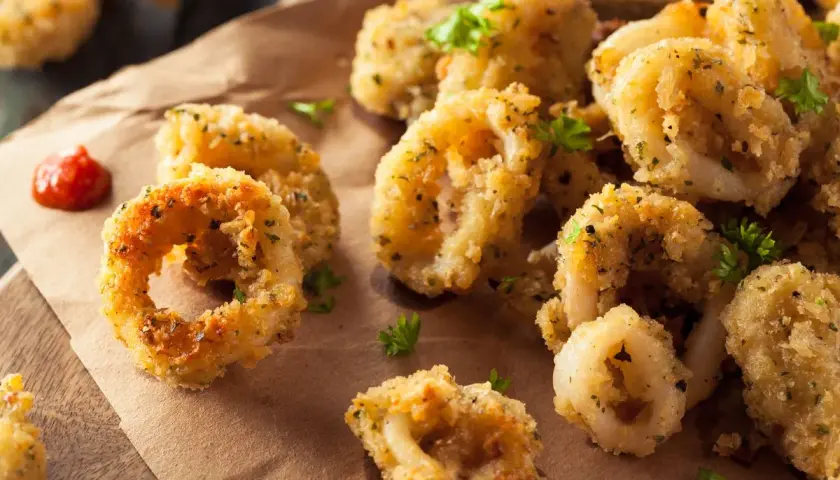 Anelli dorati e croccanti di calamari gratinati al forno, guarniti con prezzemolo fresco e accompagnati da una salsa rossa.