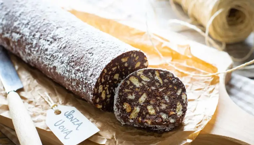 Salame al cioccolato senza uova, cosparso di zucchero a velo, con una fetta tagliata che mostra i biscotti sbriciolati.