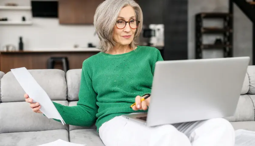 Femme âgée souriante, assise sur un canapé, travaillant sur son ordinateur portable et consultant des documents. Elle semble gérer son **apa à domicile montant**.