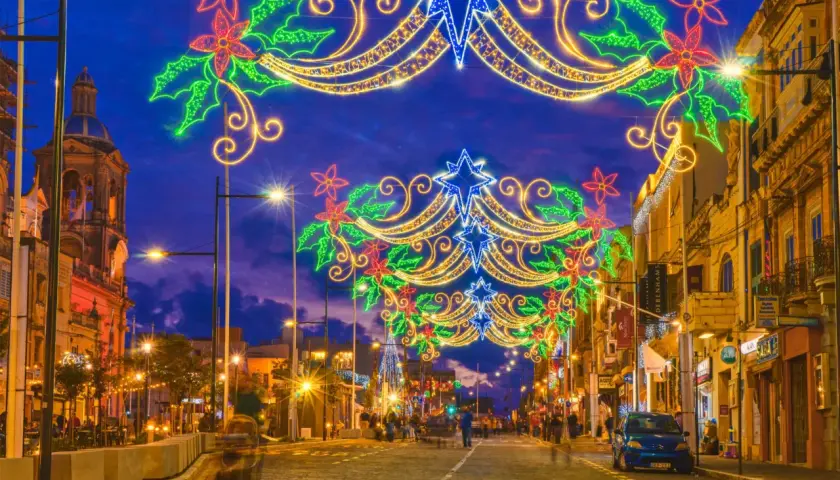 Malta w grudniu rozświetlona świątecznymi iluminacjami. Ulica ozdobiona girlandami z gwiazd i kwiatów, w tle kościół i sklepy.