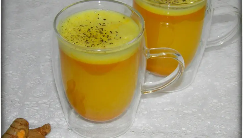 Zwei Gläser mit leuchtend gelbem Kurkuma-Wasser, bestreut mit schwarzem Pfeffer. Daneben liegen frische Kurkumawurzeln. Ein einfaches Rezept für Wohlbefinden.