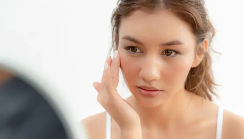 Une jeune femme examine son visage, préoccupée par la sécheresse cutanée. Sa peau semble tendue et manque d'hydratation.