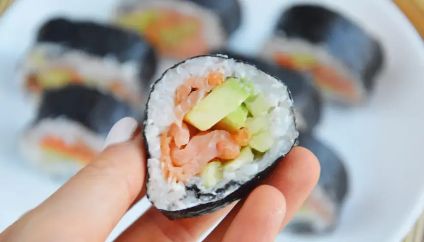 Ręka trzyma sushi z łososiem wędzonym na gorąco, awokado i ogórkiem.