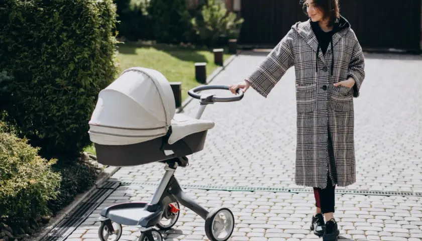 Kobieta w płaszczu prowadzi spacerówkę kubelkową po brukowanej ścieżce, wśród zieleni.
