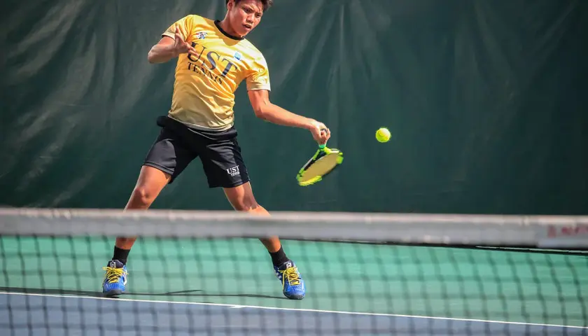 Młody tenisista w żółtej koszulce UST TENNIS uderza piłkę, walcząc o każdy break point.