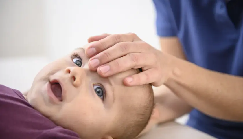 Ein Baby nach Osteopathie, wie ausgewechselt, mit weit geöffnetem Mund und großen Augen, während eine Hand sanft seine Stirn berührt.