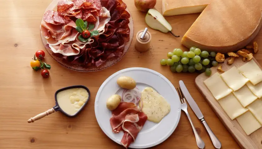 Une table dressée pour une abondance raclette : charcuteries, fromages, pommes de terre, raisins et poêlon de fromage fondu.