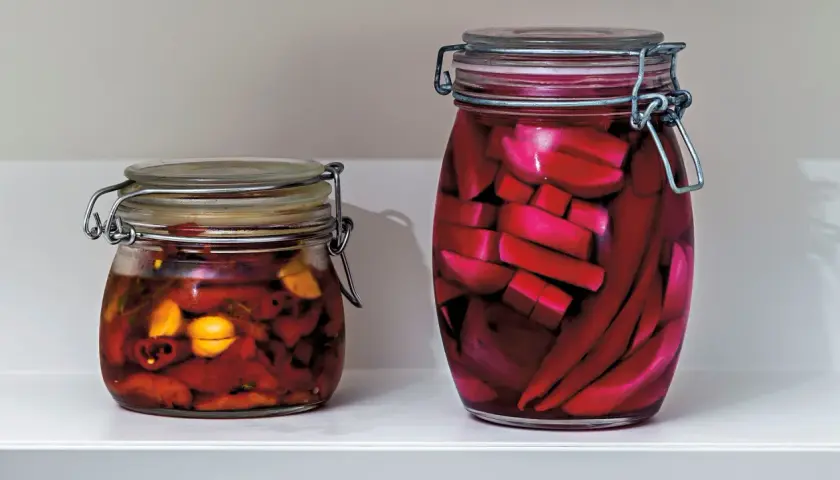 Två glasburkar med inlagd paprika och andra grönsaker. Den större burken innehåller röda, avlånga bitar, troligen inlagd paprika.