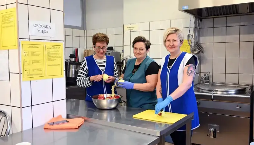 Trzy kobiety w fartuchach obierają jabłka w szkolnej kuchni.