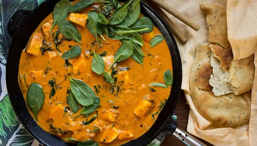En krämig palak paneer halloumi i en gjutjärnspanna, toppad med färsk spenat. Bredvid ligger naanbröd.