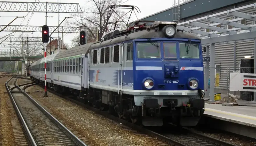 Niebieski pociąg TLK EU07-067 z wagonami PKP Intercity stoi na torach obok peronu.