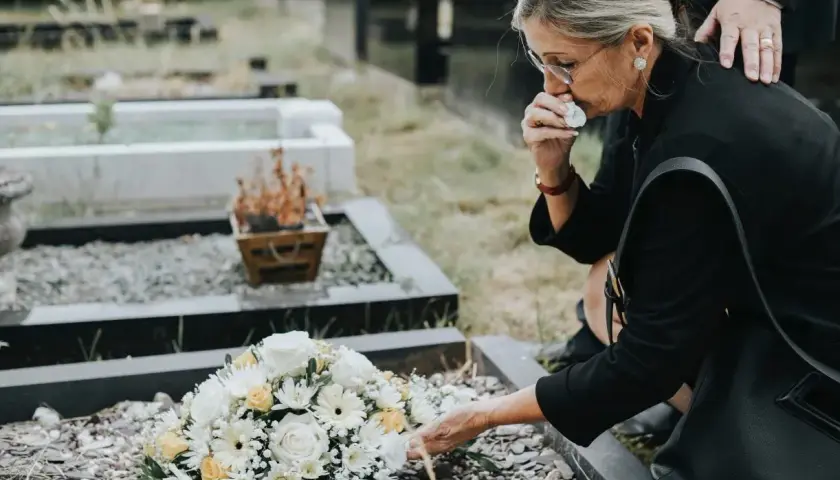 Kobieta w żałobie klęczy przy grobie, ocierając łzy. Zastanawia się, ile wynosi zasiłek pogrzebowy, by móc godnie pożegnać bliską osobę.