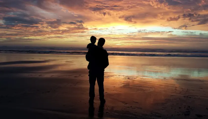 Silhouette di un padre che porta in braccio il figlio sulla spiaggia al tramonto. Un momento di gioia che celebra la festa del papà nel mondo.