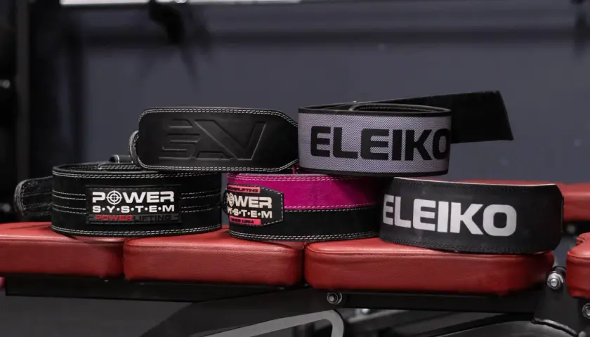 Kilka pasów do dźwigania, w tym marki Eleiko i Power System, leży na czerwonej ławce treningowej.