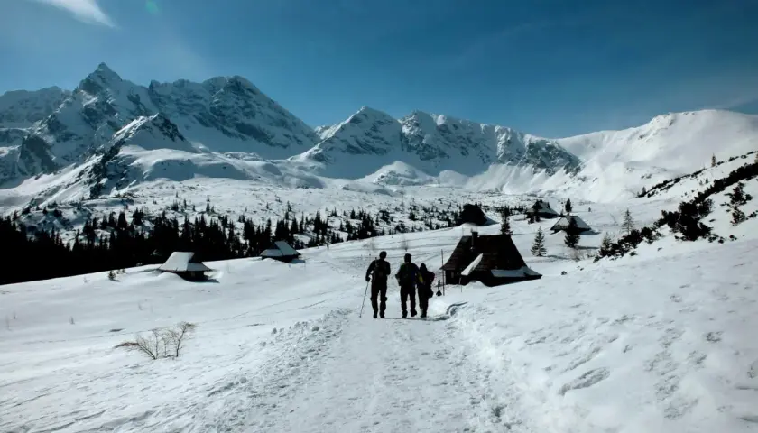 Trzej turyści idą szlakiem przez zaśnieżone Tatry zimą dla średniozaawansowanych, mijając góralskie chatki.