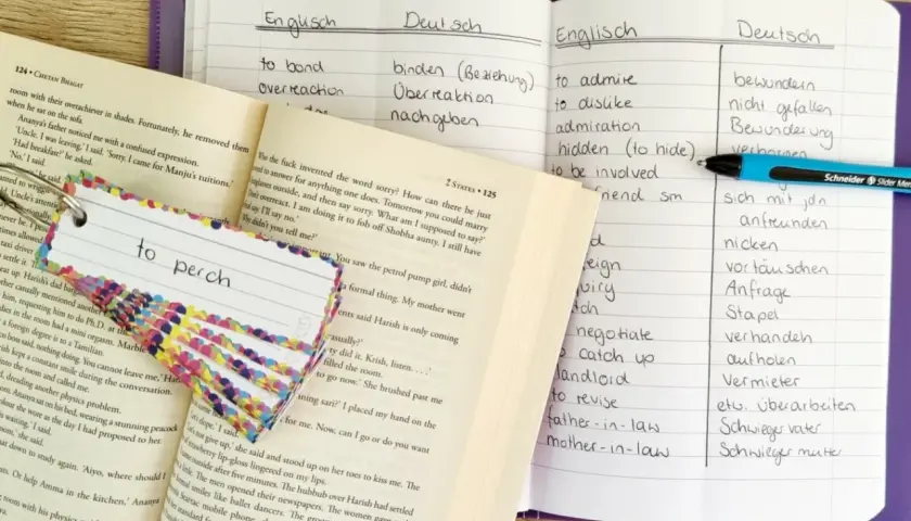 Ein offenes digitales Vokabelheft mit englischen und deutschen Wörtern, daneben ein Buch und Karteikarten zum Lernen.