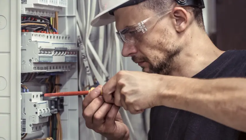 Technik elektryk w okularach ochronnych i kasku precyzyjnie dokręca śrubę w skrzynce rozdzielczej.