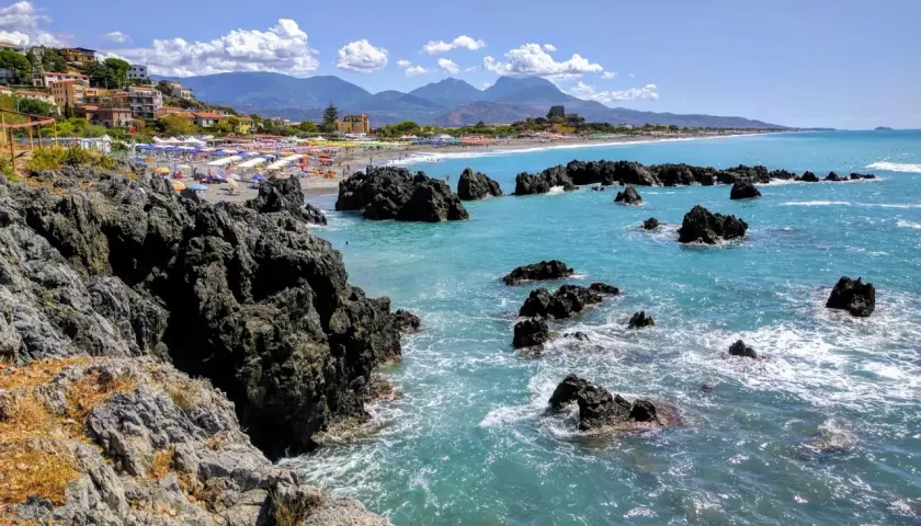 Vue d'une plage animée avec des parasols colorés, des rochers sombres et une mer turquoise. La **scalea** de la côte italienne offre un paysage pittoresque.