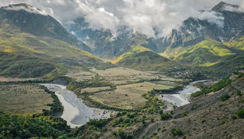 Ein malerischer Fluss schlängelt sich durch ein grünes Tal in Albanien, umgeben von bewaldeten Bergen unter dramatischen Wolken.