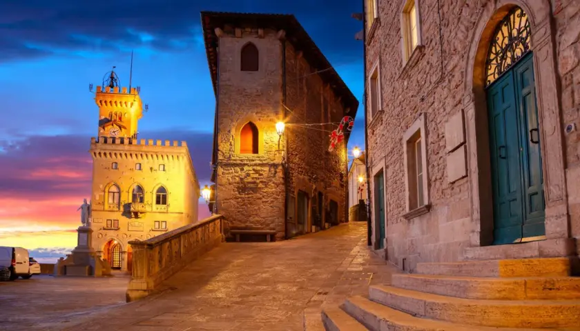 Wieczorna panorama San Marino, z wieżą i kamiennymi budynkami oświetlonymi ciepłym światłem. Niebo mieni się odcieniami różu i błękitu.