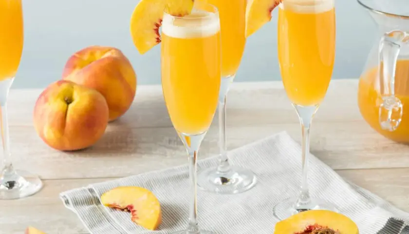 Tre glas med en gyllene drink, garnerade med persikoklyftor. Persikor och en karaff med drinken syns också.