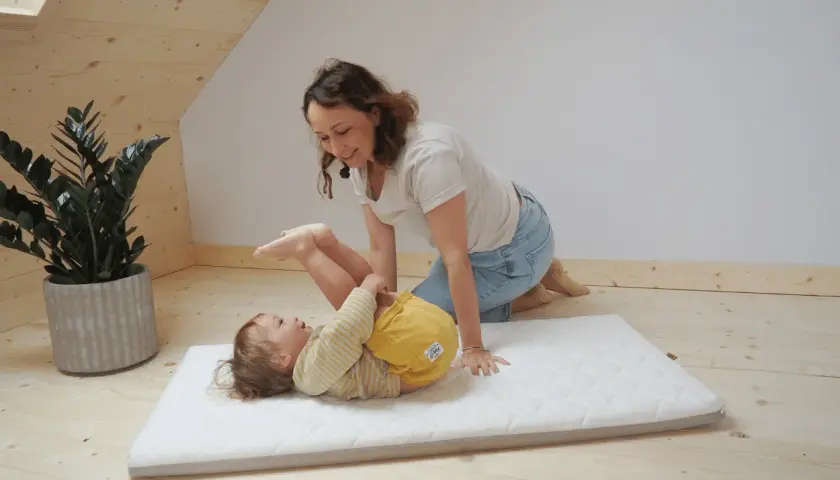 Mama bawi się z dzieckiem na materacu. Czy do łóżeczka turystycznego potrzebny jest materac? Tak, dla komfortu malucha.