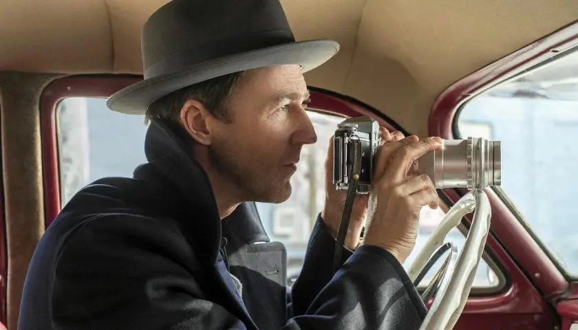 Edward Norton w filmach, w kapeluszu i płaszczu, siedzi za kierownicą samochodu i robi zdjęcie starym aparatem.