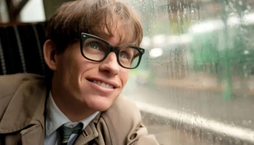 Junger Stephen Hawking mit Brille blickt lächelnd aus einem Fenster, das von Regentropfen bedeckt ist.
