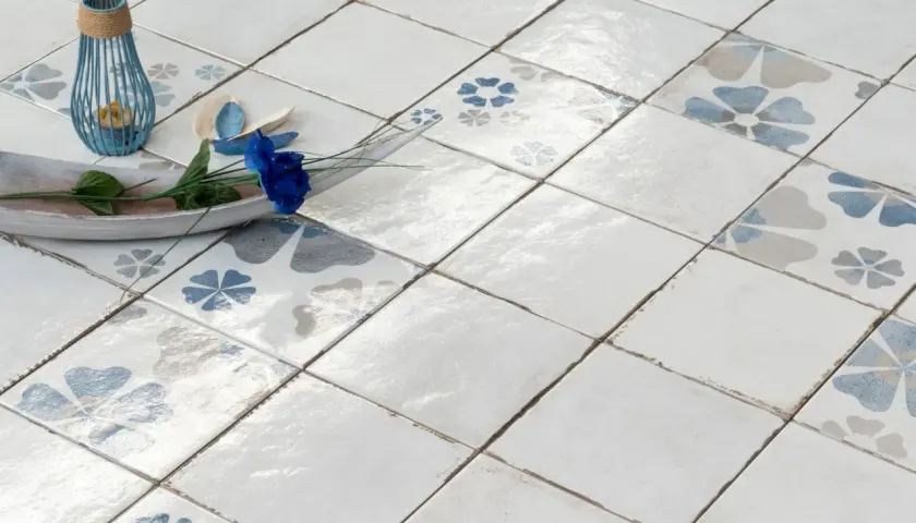 Pavimento con piastrelle quadrate, alcune decorate con fiori blu e grigi, altre bianche. Un vassoio in legno con un fiore blu e un portacandele blu completano la scena, mostrando la differenza tra mattonella e piastrella.