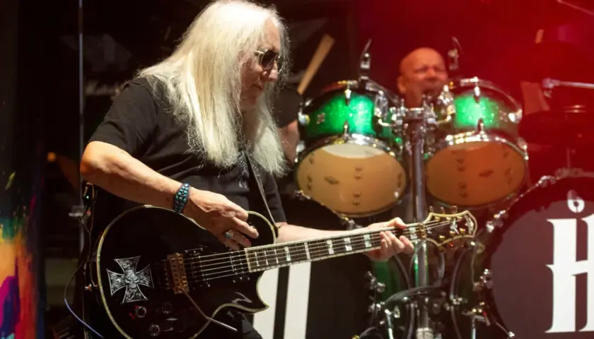 Uriah Heep członkowie na scenie. Gitarzysta z długimi blond włosami gra na czarnej gitarze z symbolem krzyża. W tle perkusista i zielone bębny.