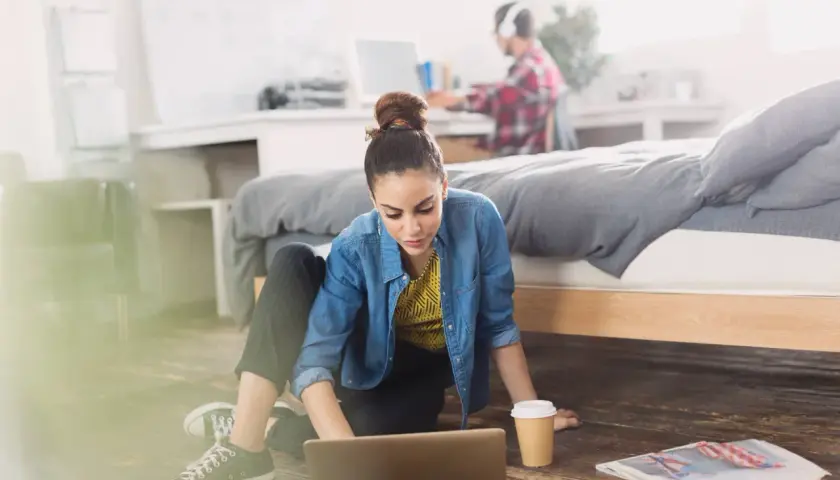 Młoda kobieta pracuje na laptopie na podłodze w pokoju. Obok niej kubek kawy i gazeta. W tle mężczyzna w słuchawkach. Zastanawia się, ile osób może mieszkać w wynajmowanym mieszkaniu.