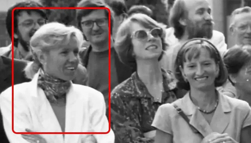 Brigitte Macron młoda, w białej marynarce i z apaszką na szyi, uśmiecha się do aparatu. W tle widać innych ludzi.