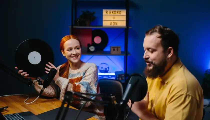 Coppia sorridente registra podcast in inglese interessanti, lei tiene un vinile, lui parla al microfono.