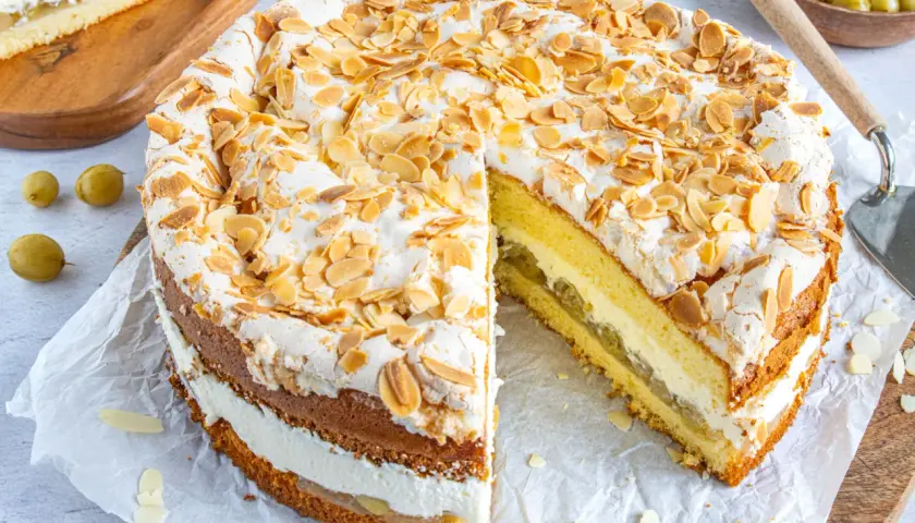 Ein köstlicher Stachelbeer Baiser Kuchen mit Biskuitböden, Sahne und Mandelsplittern. Ein Stück ist bereits angeschnitten.