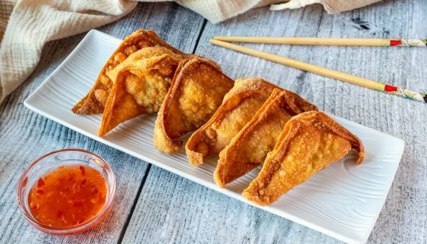 Crujientes wan tun frito dorados en una bandeja blanca, acompañados de salsa agridulce y palillos.