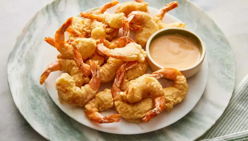 A plate piled high with golden-brown shrimp tempura, served with a small bowl of creamy dipping sauce.