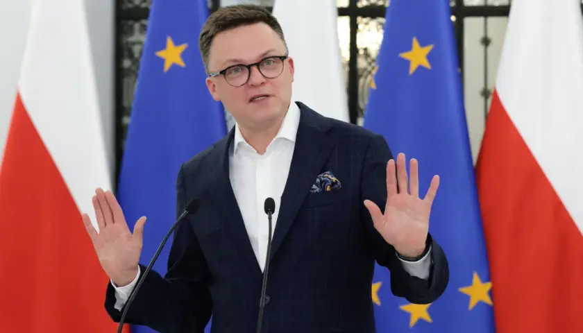Szymon Hołownia w garniturze przemawia przed flagami Polski i UE.