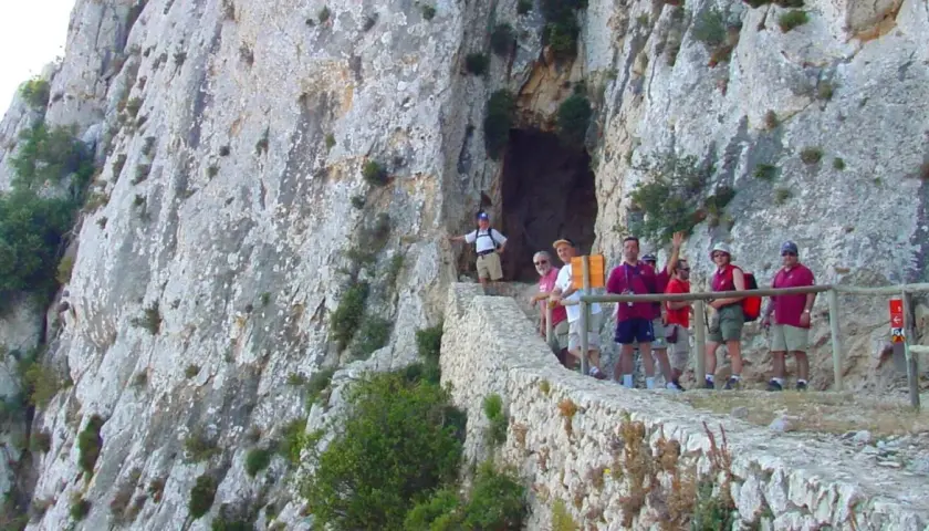 Grupo de excursionistas en el sendero del Peñón de Calpe, con una cueva a la izquierda y vistas rocosas.