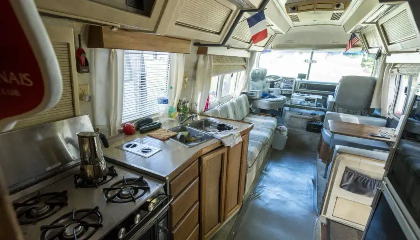 Vue de l'intérieur d'un camping-car américain : cuisine avec cuisinière, évier, et banquette. Drapeaux français et américain visibles.
