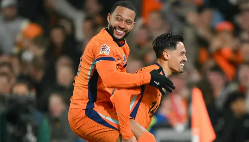 Dwóch piłkarzy Holandii w pomarańczowych strojach świętuje bramkę. Jeden z nich, Memphis Depay, niesie kolegę na plecach, obaj uśmiechnięci.