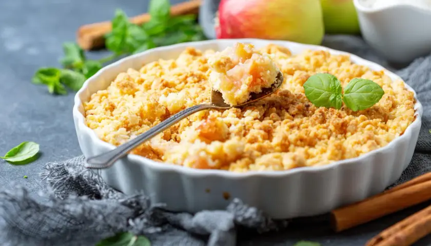 Un délicieux crumble pomme doré, garni de feuilles de menthe fraîche. Des pommes rouges et vertes sont visibles en arrière-plan.